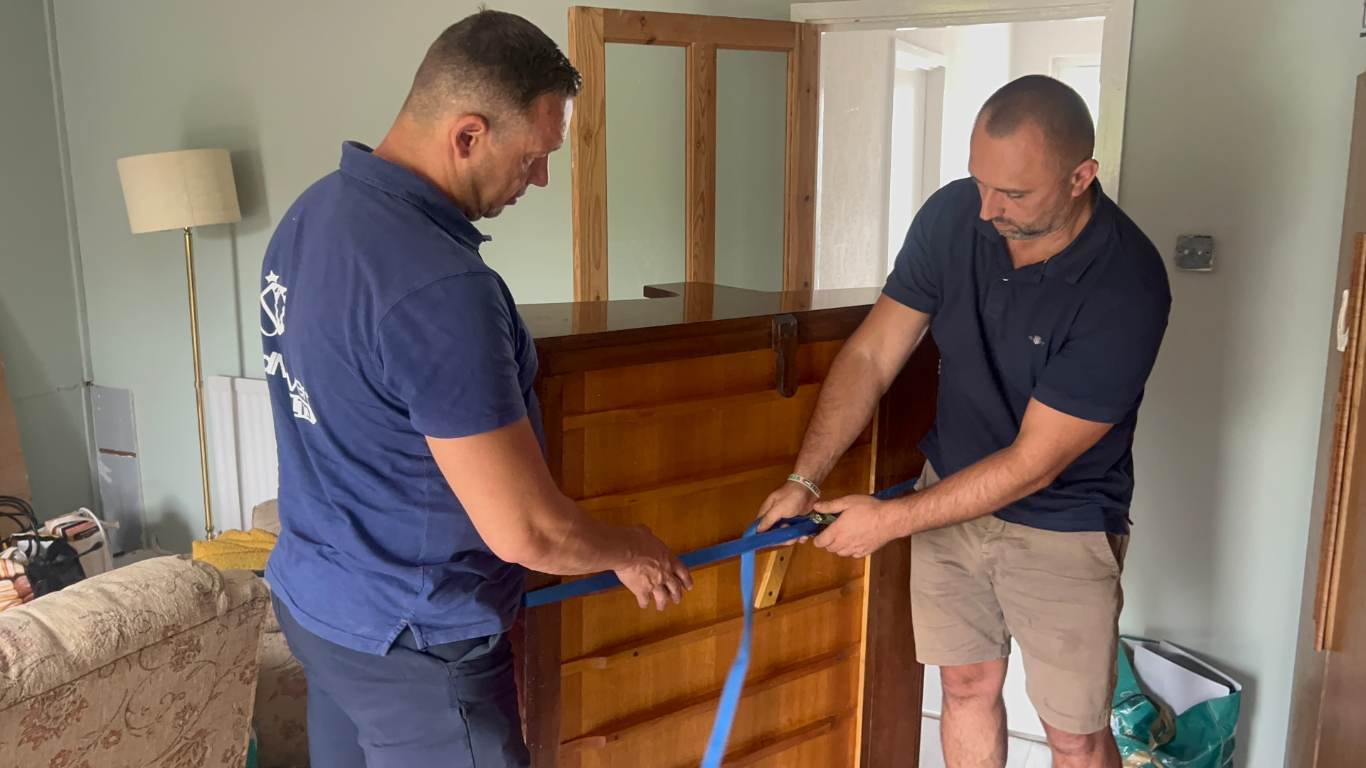 Two Dimond Movers piano specialists fitting heavy-duty ratchet straps to an antique upright piano inside a living room