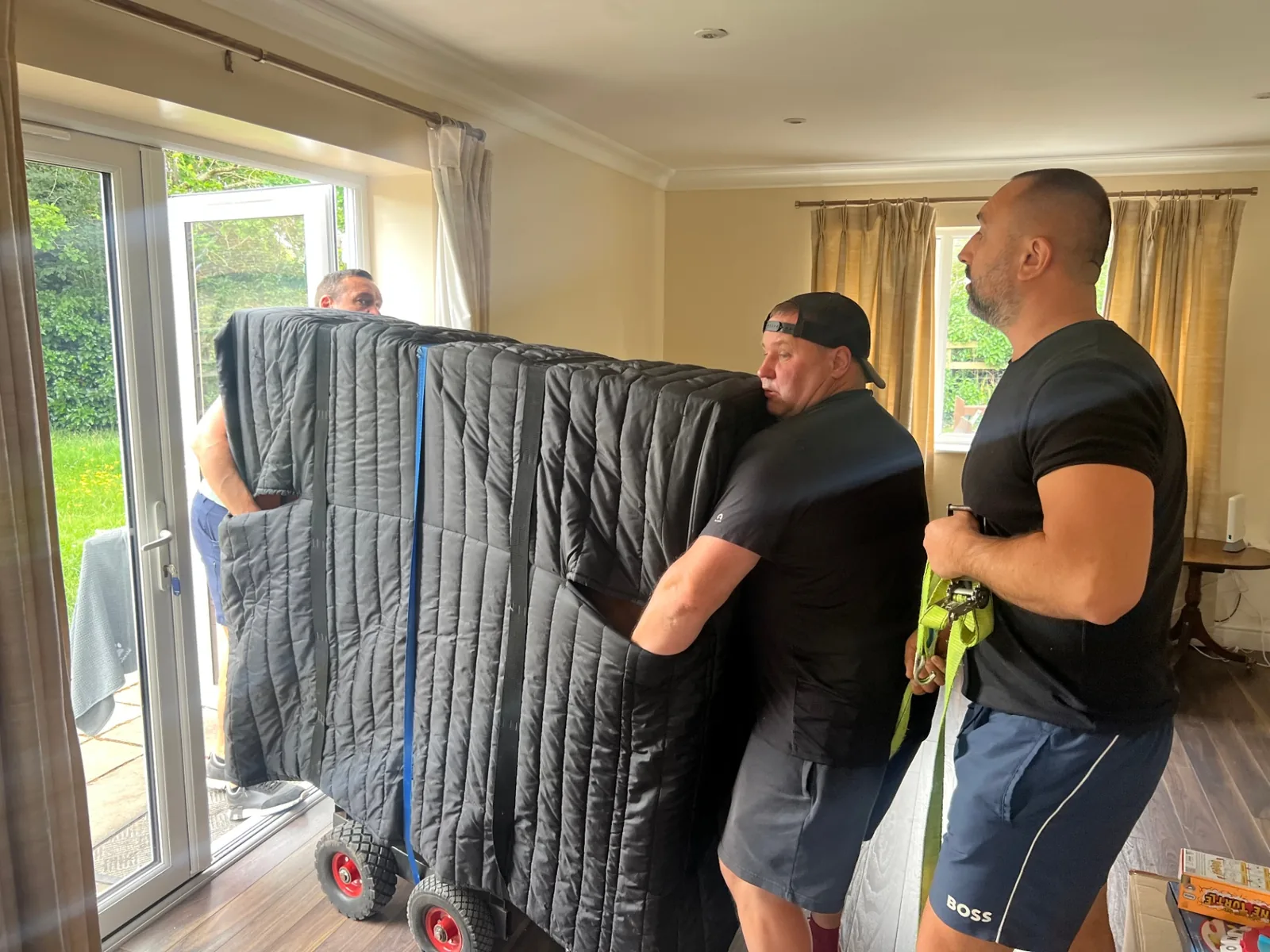 Dimond Movers piano team carefully wheeling a fully wrapped upright piano out through a UK home's patio doors